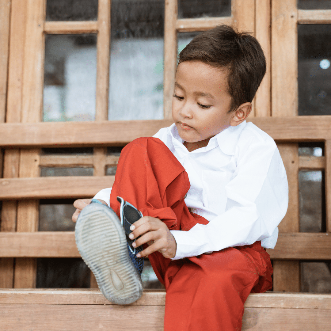 Image: A young boy in a white shirt and red pants sitting on a bench putting on his boots.