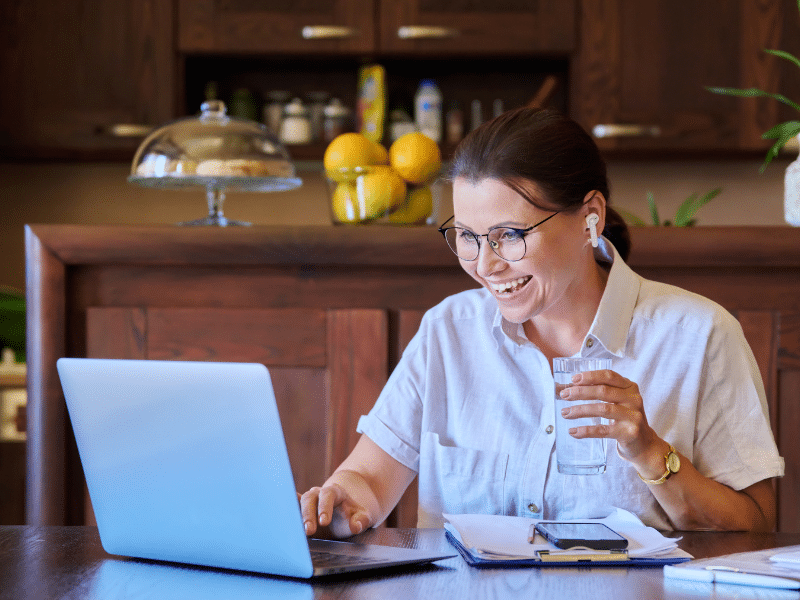A mother with glasses and a button-up shirt smiles as she uses a laptop. She seems engaged and cheerful, with wireless earphones in her ears and a glass of water in her hand. She is accessing positive behaviour support therapy coaching. The setting appears to be a home kitchen, with fruit and kitchenware in the background.
