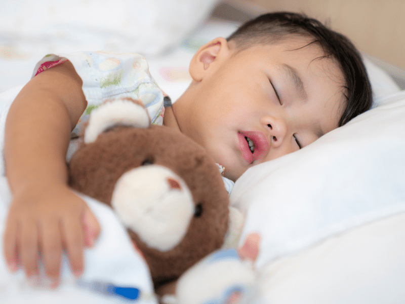 A young child is in a peaceful slumber, cuddling a brown and white teddy bear. The child is dressed in a light, patterned onesie, suggesting it's time for a nap or bedtime. The environment is calm, with soft bedding ensuring a comfortable rest.
