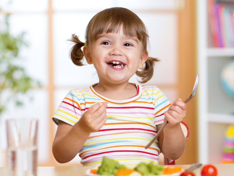 A joyful toddler with pigtails is excitedly eating, holding a spoon in one hand and a bite of food in the other. She's wearing a colorful striped t-shirt, and in front of her is a plate full of healthy foods like broccoli and carrots, indicating a positive mealtime experience. The background is bright and homey, likely a dining area.
