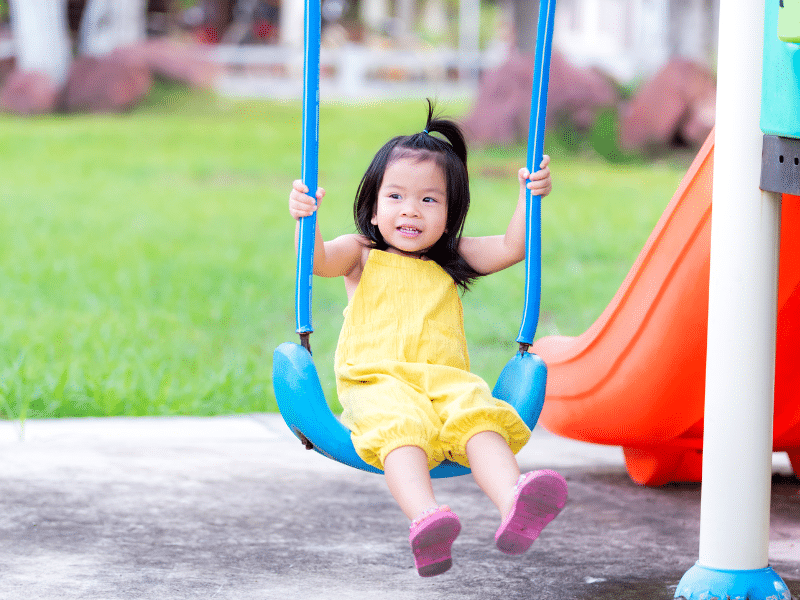 Image: A young girl in a yellow romper on a blue swing at the playground.